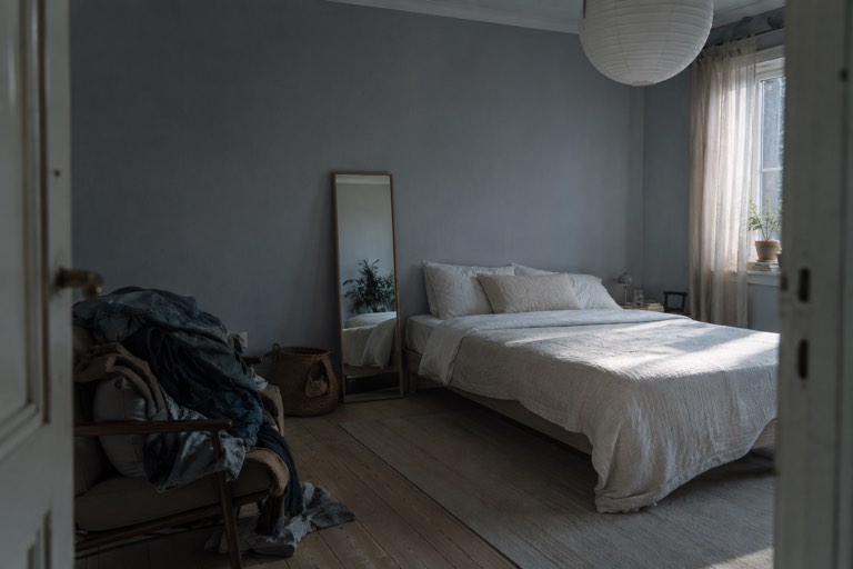 A bedroom seen from the doorway, a freshly made bed in daylight on the right and an armchair piled with clothes in shadow on the left