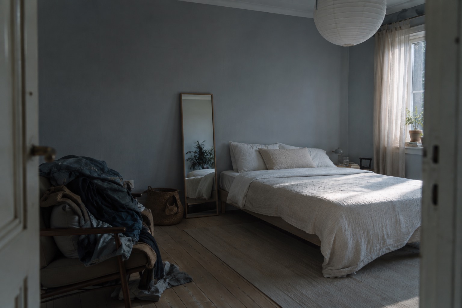 A bedroom seen from the doorway, a freshly made bed in daylight on the right and an armchair piled with clothes in shadow on the left
